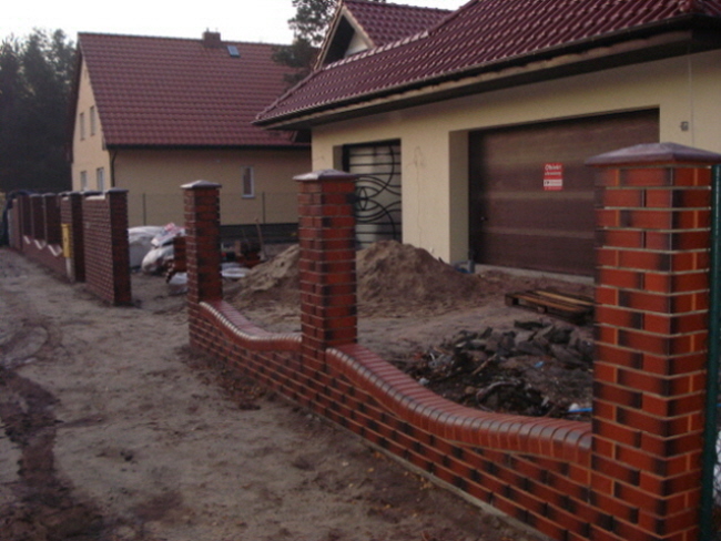 Wznoszone ogrodzenie z cegły klinkierowej z falistym fragmentem, widoczne słupki i przęsła w trakcie budowy, na tle dom z garażem i sterta piasku.