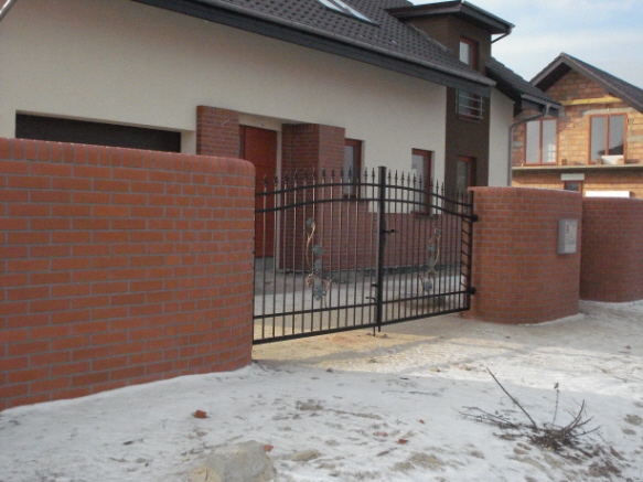 Nowoczesne ogrodzenie z czarnych prętów z dekoracyjnymi elementami, osadzone na murkach z cegły klinkierowej, na tle domu z jasną elewacją i resztkami śniegu.