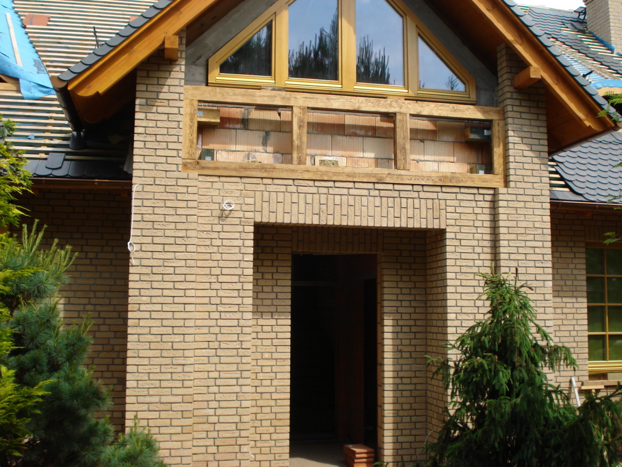 Elewacja budynku w trakcie budowy, z widoczną klinkierową cegłą ręcznie formowaną, drewnianą stolarką okienną i fragmentem dachu pokrytego dachówką. Widoczne elementy konstrukcyjne i brakujące...