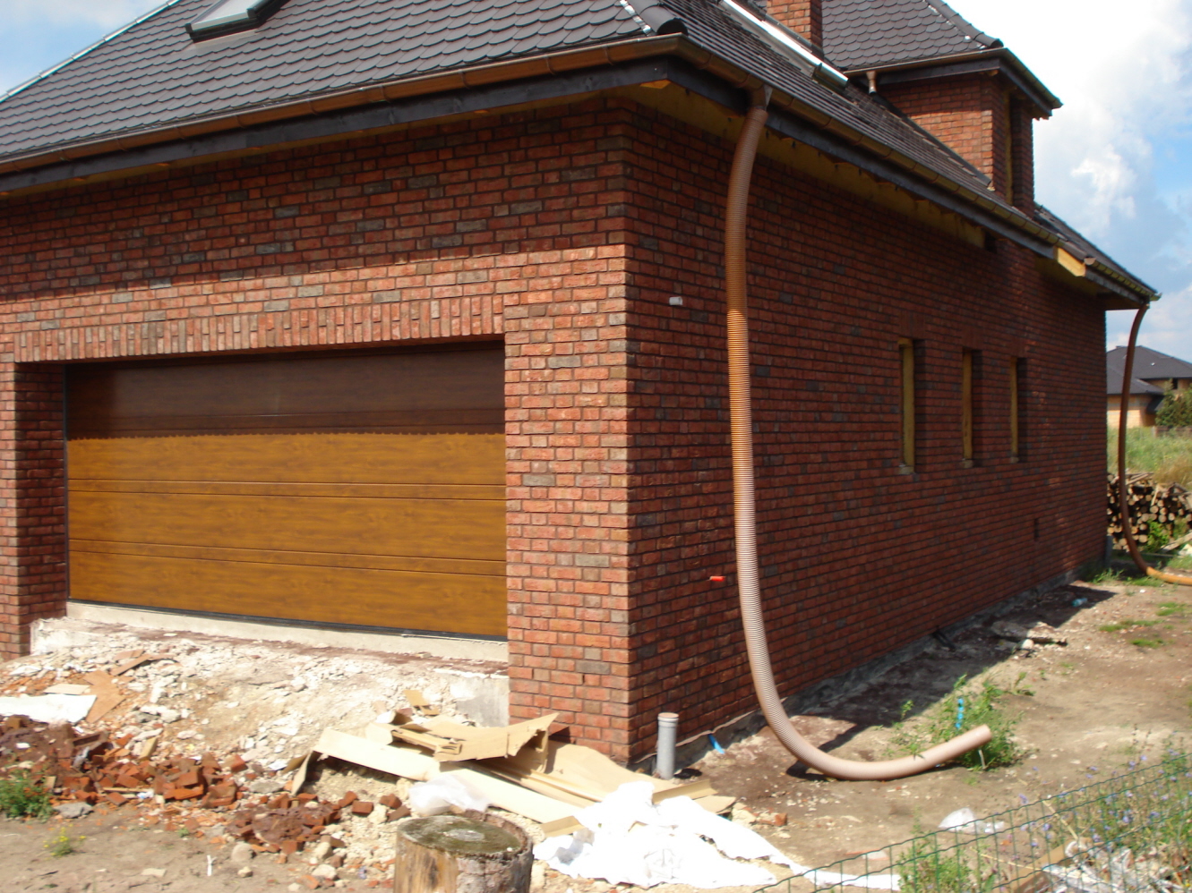 Elewacja budynku z czerwonej cegły klinkierowej z brązową bramą garażową i rurą spustową. Widoczne ślady prac budowlanych wokół budynku.