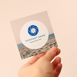 EMPIK FOTO SPÓŁKA Z OGRANICZONĄ ODPOWIEDZIALNOŚCIĄ - Dłoń trzyma wizytówkę z motywem plaży i logo firmy. Wizytówka ma okrągłe logo z niebieskim symbolem przysłony i nazwą firmy. Tło wizytówki imituje piasek i morze.