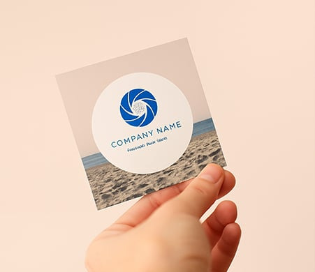 Dłoń trzyma wizytówkę z motywem plaży i logo firmy. Wizytówka ma okrągłe logo z niebieskim symbolem przysłony i nazwą firmy. Tło wizytówki imituje piasek i morze.