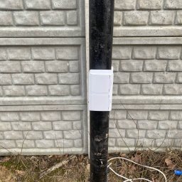 SUE - Biała skrzynka elektryczna na czarnej rurze przy betonowym ogrodzeniu w Żyrardowie. Widoczne kable i ziemia z liśćmi.