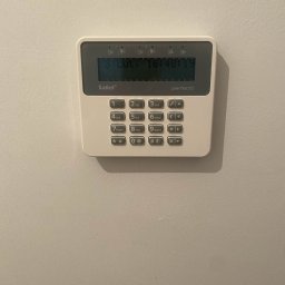 SUE - Panel sterowania alarmu Satel Perfecta na ścianie, wyświetlający datę i godzinę. Biała obudowa, szare przyciski numeryczne i funkcyjne. Widoczne logo Satel.