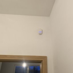 SUE - Biały czujnik alarmowy z niebieską diodą LED zamontowany na ścianie nad drewnianą futryną drzwi. Minimalistyczny design, nowoczesne zabezpieczenia.