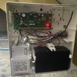 SUE - Otwarta obudowa centrali alarmowej z widoczną elektroniką, przewodami, transformatorem i akumulatorem. Czerwona dioda LED sygnalizuje działanie systemu.