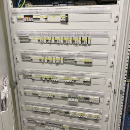 SUE - Rozdzielnica elektryczna z licznymi zabezpieczeniami i oznaczeniami, widoczne przewody i elementy sterujące. Całość w metalowej obudowie.