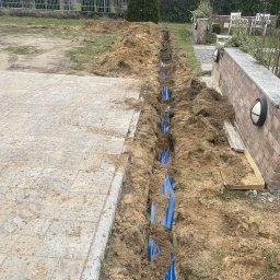 SUE - Wykop z niebieskimi rurami w ziemi, prawdopodobnie instalacja podziemna na posesji. Widoczny fragment kostki brukowej i ogrodzenie w tle.