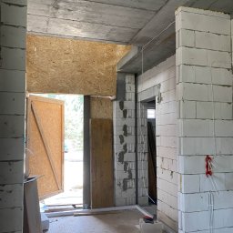 SUE - Wnętrze w trakcie budowy: surowe ściany z betonu komórkowego, widoczne przewody elektryczne, prowizoryczne drzwi z płyty OSB. Stan surowy zamknięty.