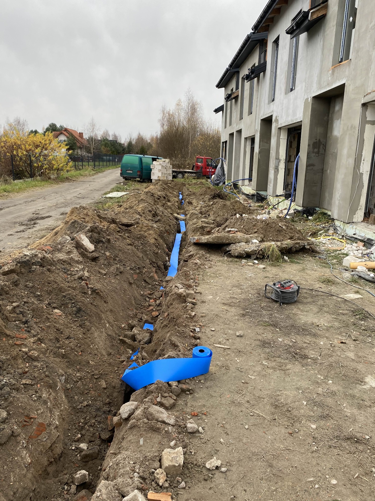 Wykop z niebieską rurą kablową na posesji z budynkiem w stanie surowym, widoczny bęben kablowy i pojazdy w tle. Etap prac instalacyjnych.
