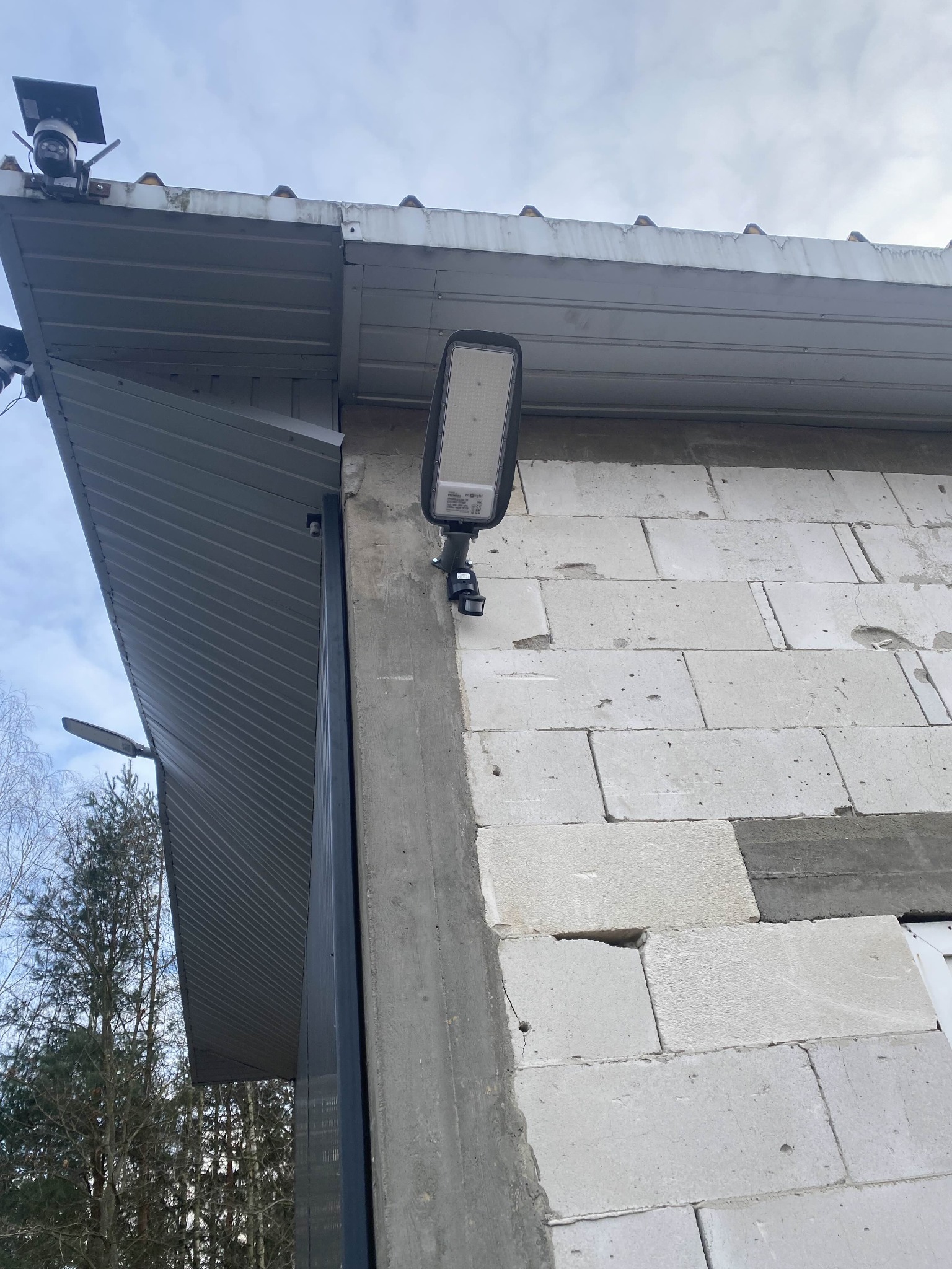 Zewnętrzna lampa LED zamontowana na ścianie budynku z betonu komórkowego, widoczna kamera monitoringu na dachu. Ujęcie z dołu, pochmurne niebo w tle.
