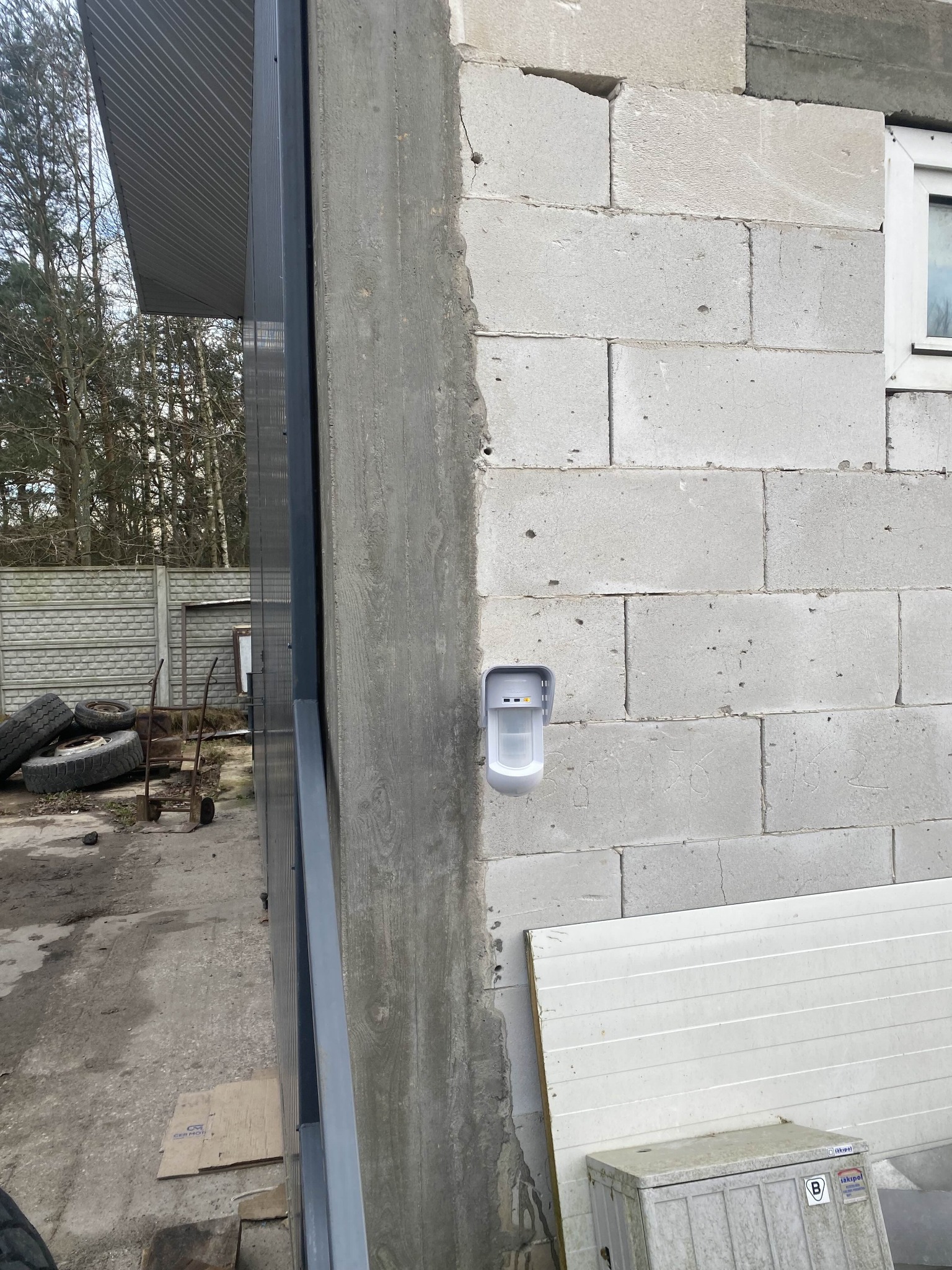 Biały czujnik ruchu zamontowany na szarej ścianie z betonu komórkowego, obok fragmentu metalowej konstrukcji i okna. Widoczne elementy otoczenia.