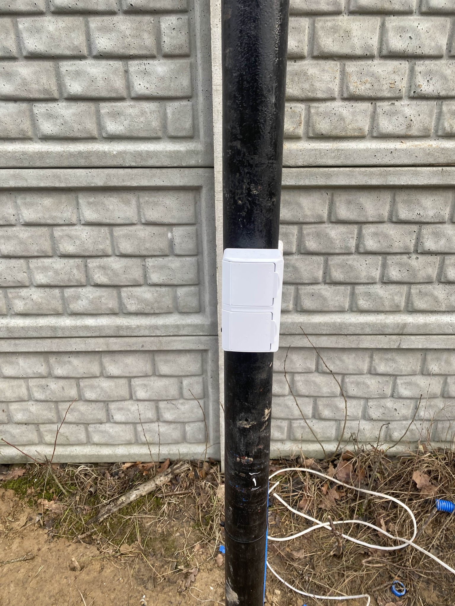 Biała skrzynka elektryczna na czarnej rurze przy betonowym ogrodzeniu w Żyrardowie. Widoczne kable i ziemia z liśćmi.