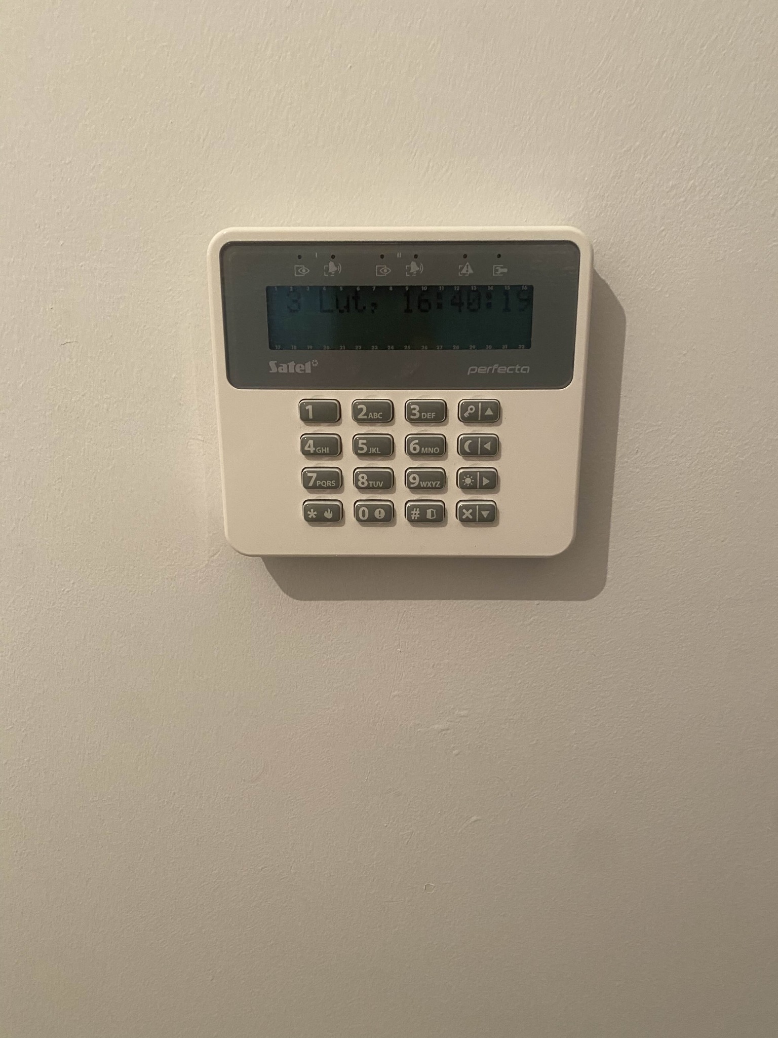 Panel sterowania alarmu Satel Perfecta na ścianie, wyświetlający datę i godzinę. Biała obudowa, szare przyciski numeryczne i funkcyjne. Widoczne logo Satel.