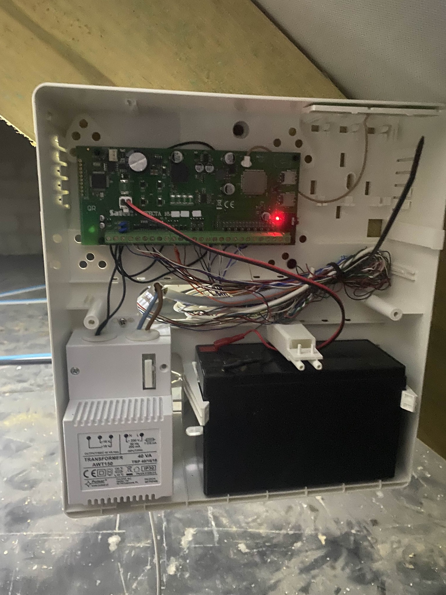 Otwarta obudowa centrali alarmowej z widoczną elektroniką, przewodami, transformatorem i akumulatorem. Czerwona dioda LED sygnalizuje działanie systemu.