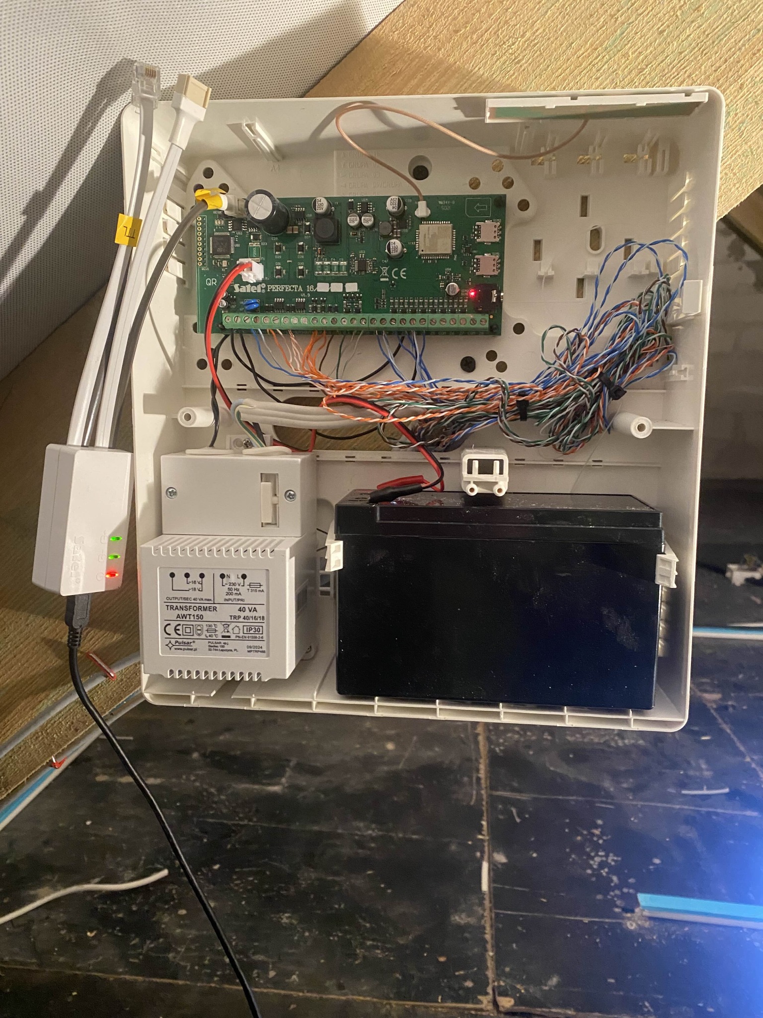 Centrala alarmowa Satel Perfecta 16 z akumulatorem, transformatorem Pulsar AWT150 i przewodami. Widoczne złącza, płytka drukowana i diody LED. Instalacja alarmowa w trakcie montażu.