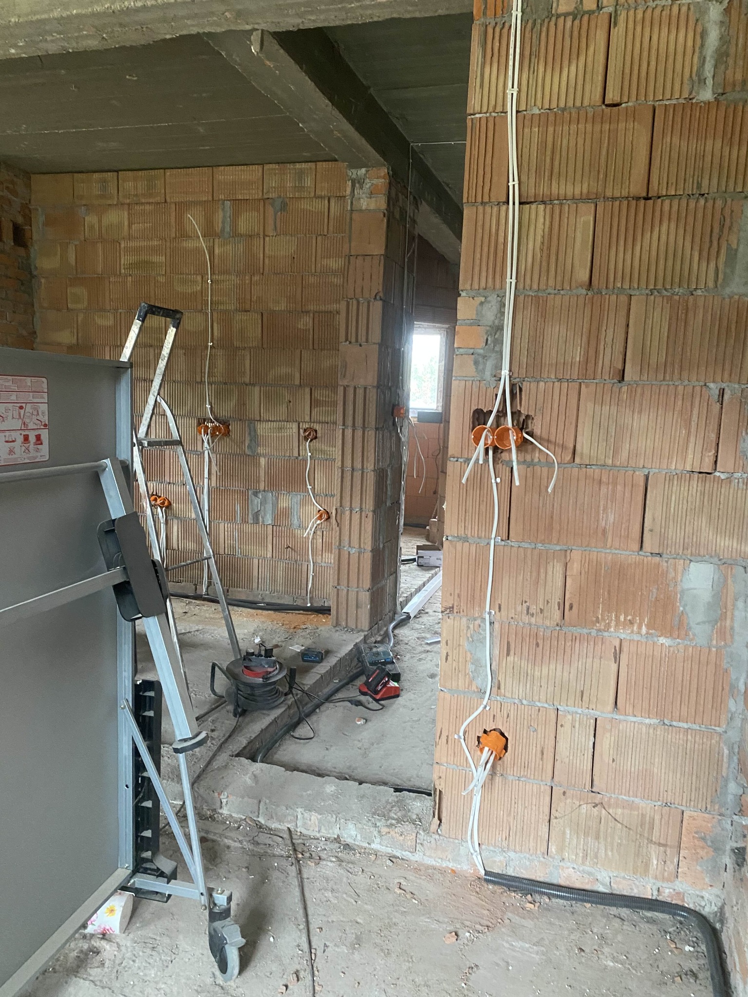 Wnętrze w trakcie prac: widoczne kable elektryczne w ścianach z cegły, puszki instalacyjne i drabina. Surowy stan wykończenia, przewody prowadzone wzdłuż ścian.