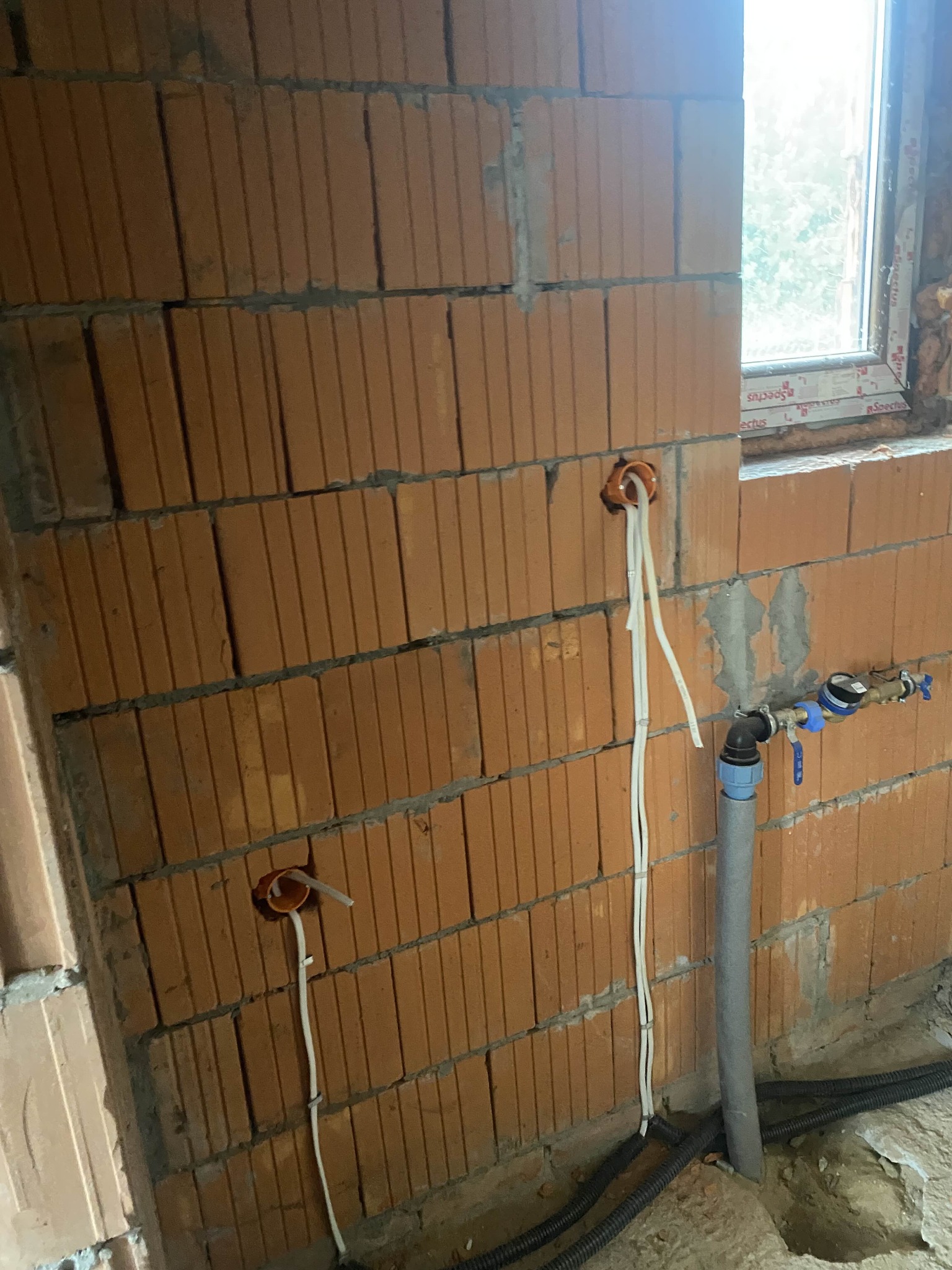Ceglana ściana z wyprowadzonymi przewodami elektrycznymi w pomarańczowych puszkach i rurą wodną z zaworem, widok z bliska na etapie prac instalacyjnych.