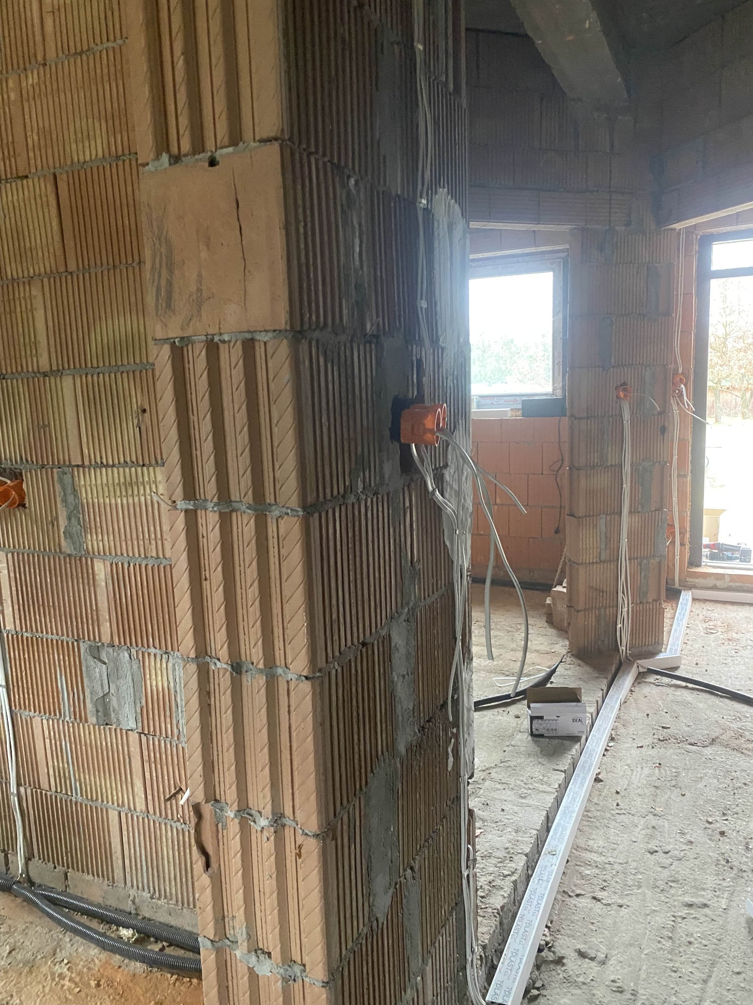 Surowy mur z instalacją elektryczną w trakcie montażu: pomarańczowe puszki i przewody. Widoczne okna i materiały budowlane. Wnętrze w stanie deweloperskim.