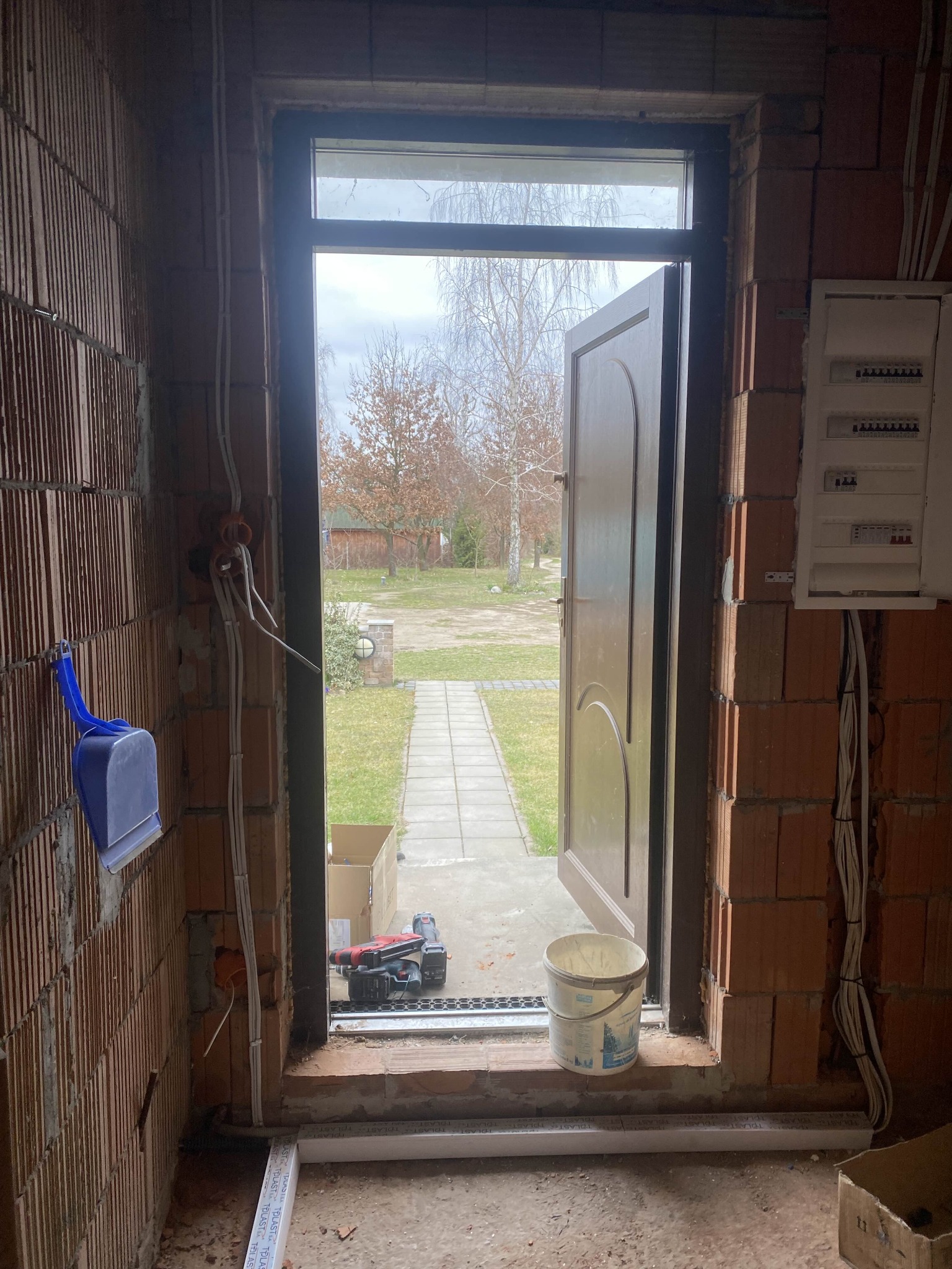 Wnętrze w trakcie remontu: widoczne przewody elektryczne na ścianie z cegły, otwarta brama na zewnątrz, narzędzia i wiadro. Etap prac instalacyjnych w nowym domu.