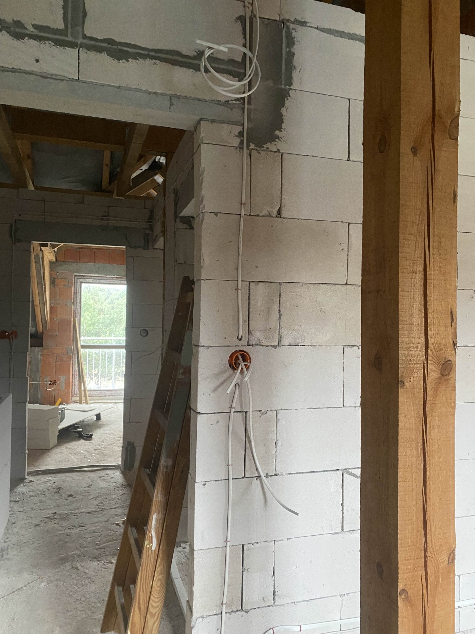 Surowy stan: Instalacja elektryczna w trakcie montażu w budynku z białych bloczków, widoczne kable i puszki, drewniana drabina oparta o ścianę, surowy stan wykończenia.