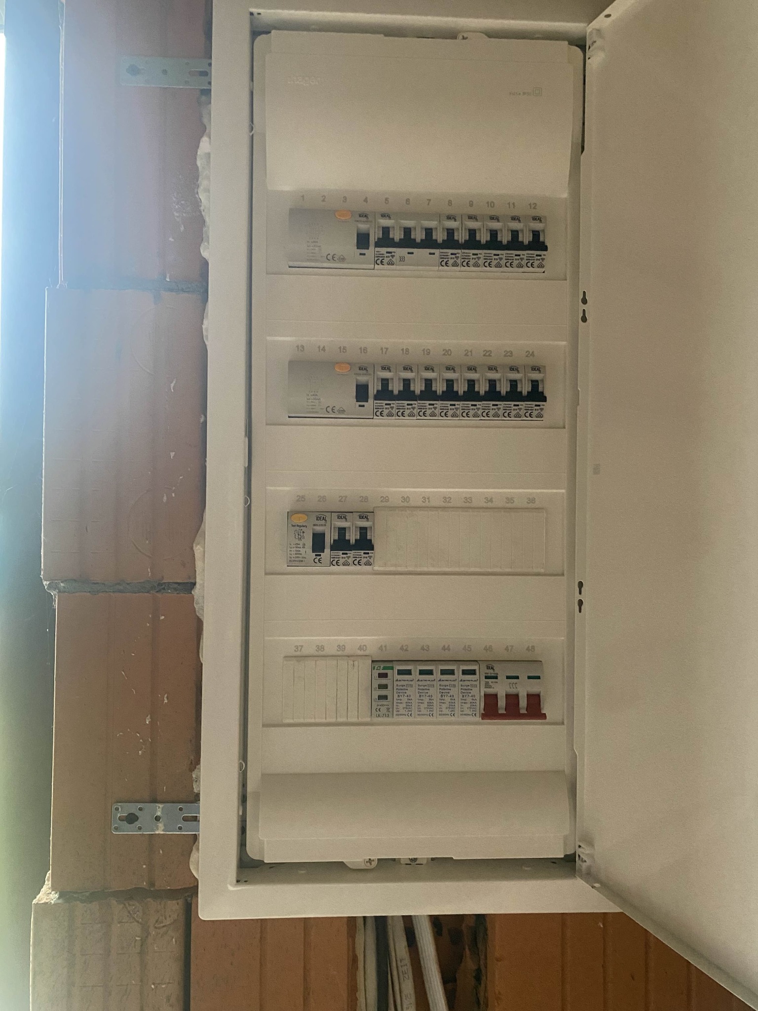 Rozdzielnica elektryczna z bezpiecznikami, zamontowana na ścianie z cegły. Widoczne numery i oznaczenia na poszczególnych elementach. Otwarta obudowa.