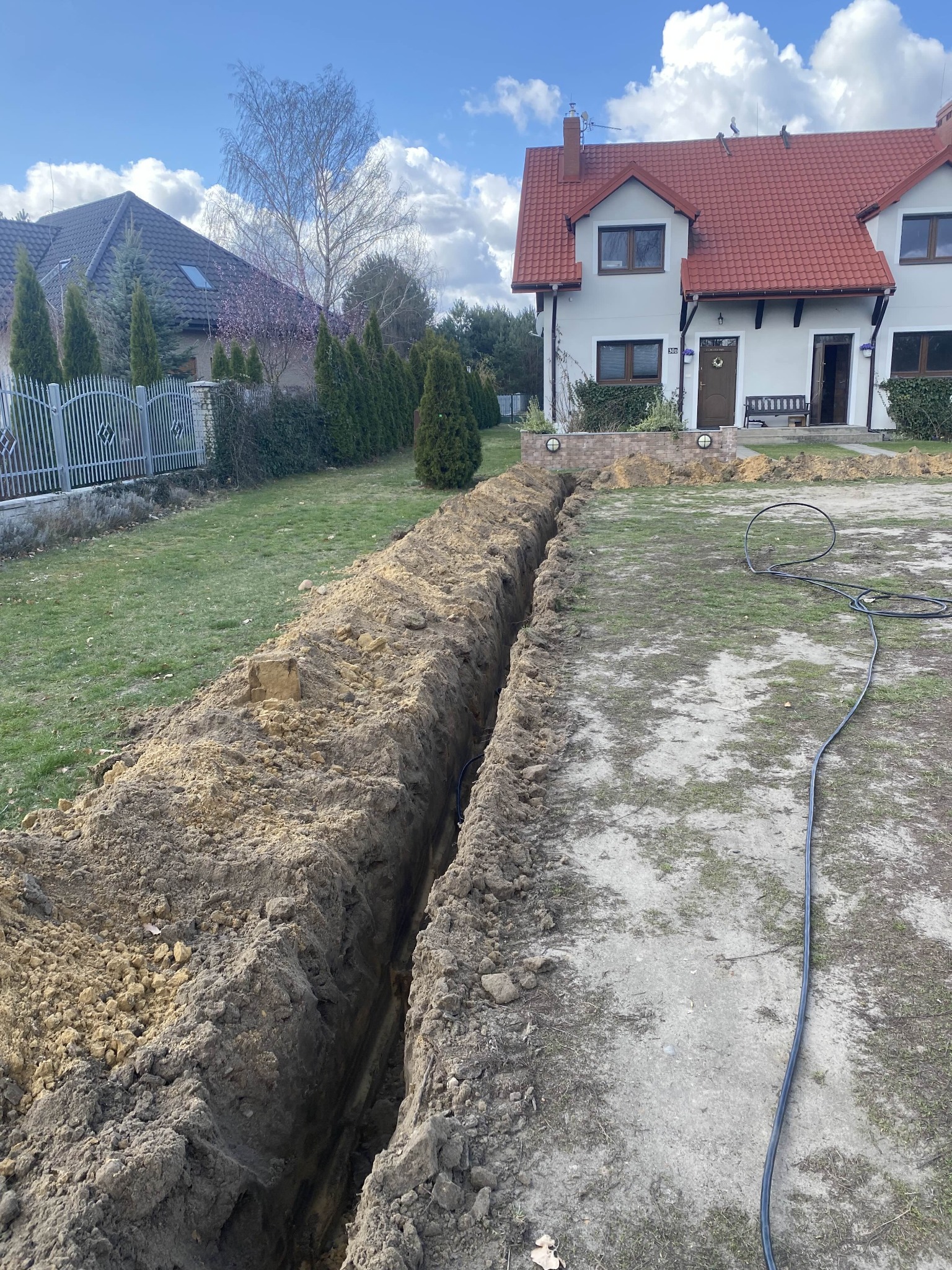 Wykop w ziemi przed domem, prawdopodobnie pod instalację elektryczną. Widoczny kabel. Domy w tle z czerwoną dachówką i ogrodzenie.