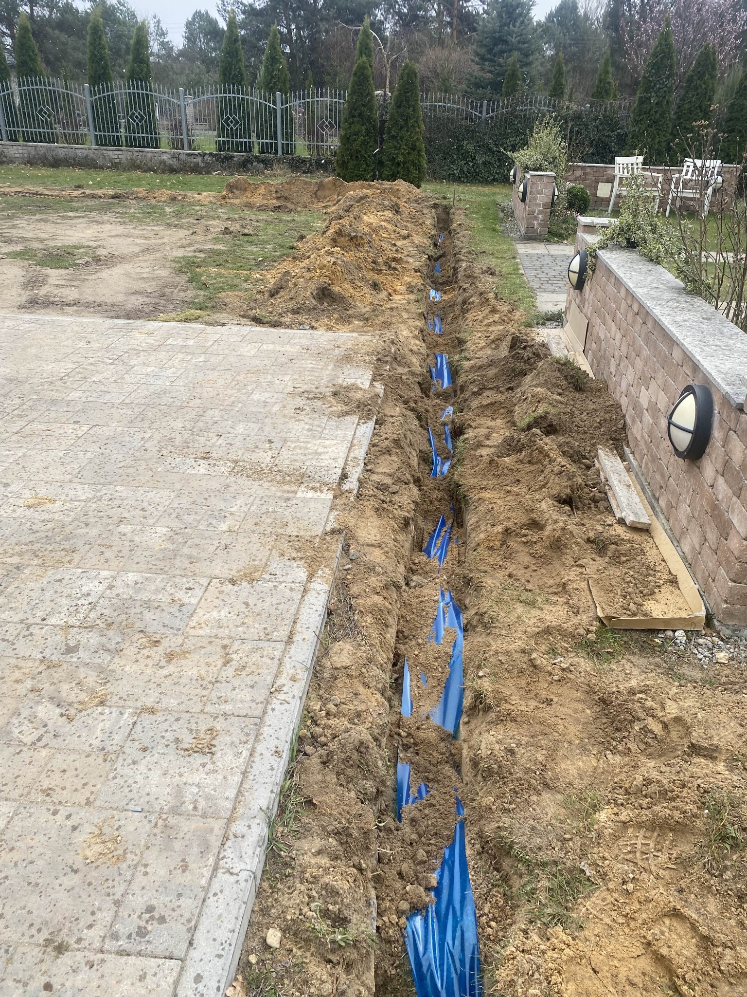 Wykop z niebieskimi rurami w ziemi, prawdopodobnie instalacja podziemna na posesji. Widoczny fragment kostki brukowej i ogrodzenie w tle.