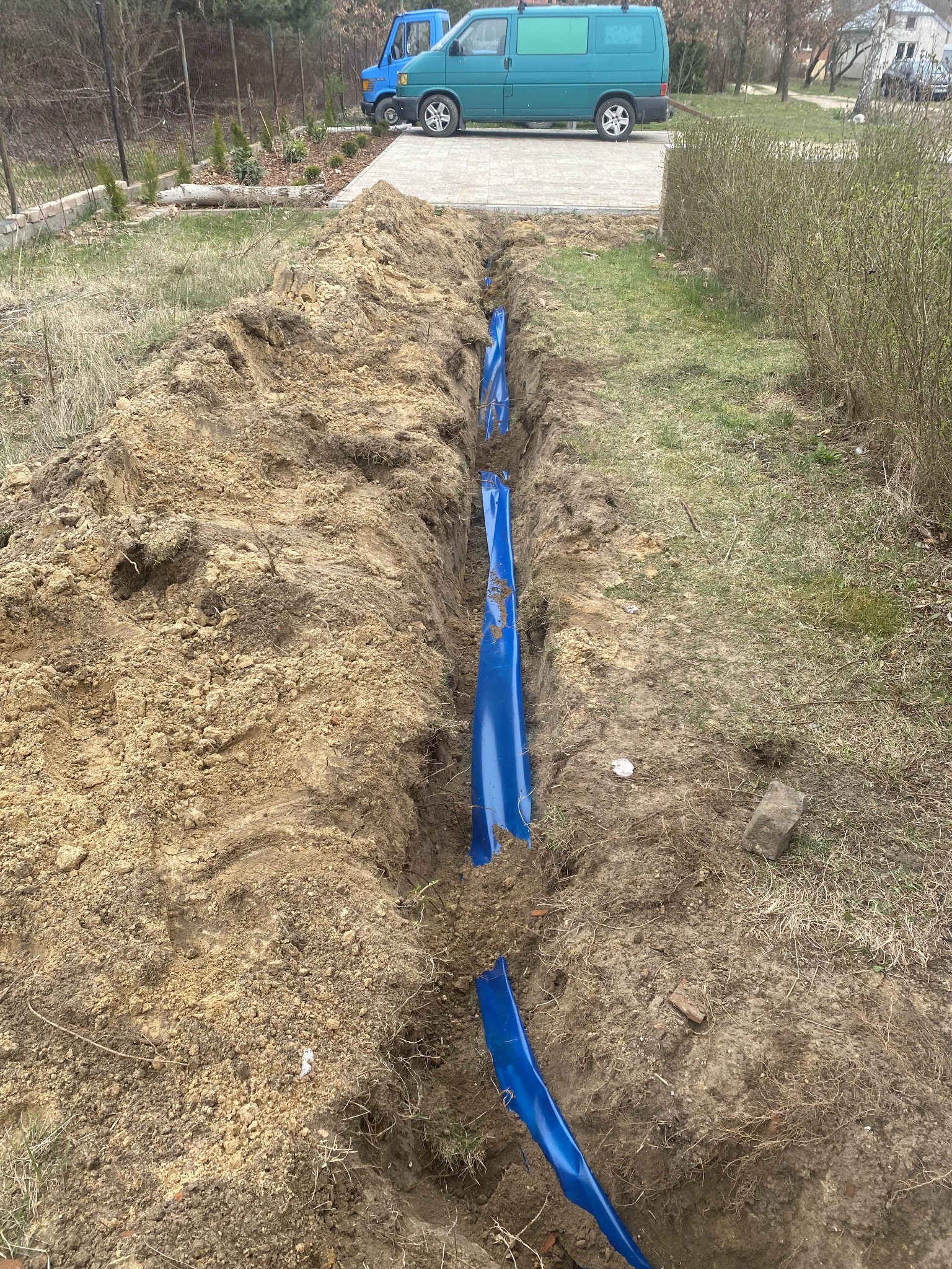 Wkopana w ziemię niebieska rura osłonowa kabla elektrycznego, widoczna w wykopie na tle trawnika i zaparkowanych samochodów dostawczych.