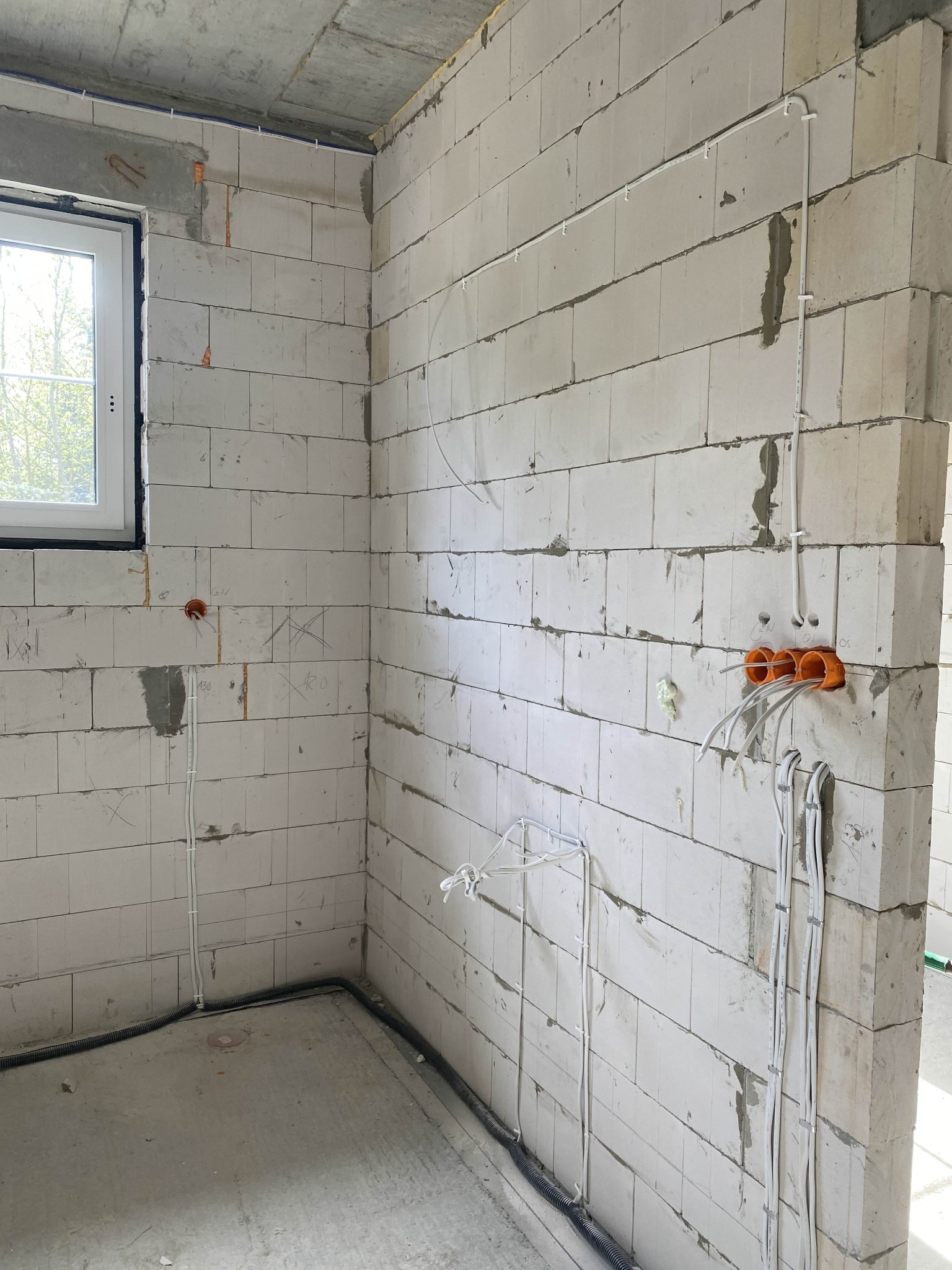 Surowy stan: instalacja elektryczna wewnątrz budynku z białych bloczków, widoczne kable, puszki i peszle. Okno z widokiem na zieleń w tle.