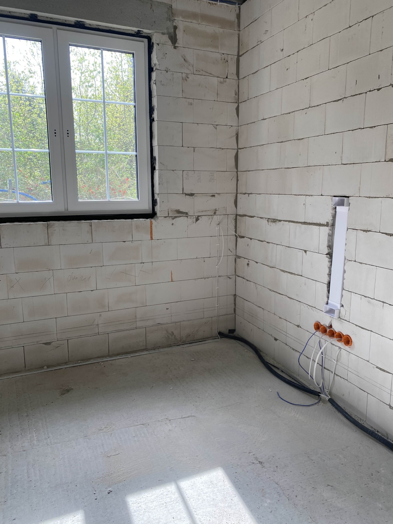 Surowy stan wnętrza z widoczną instalacją elektryczną w ścianie z betonu komórkowego. W tle okno z widokiem na zieleń. Widoczne kable i peszle.