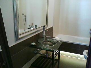 Szklana umywalka na metalowym stelażu w nowoczesnej łazience z lustrem w srebrnej ramie i białą wanną.