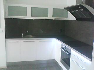 Nowoczesna kuchnia w odcieniach bieli i szarości. Białe, lakierowane szafki z matowymi przeszkleniami. Czarny panel nad blatem i podłużny okap. Minimalistyczny zlew i płyta indukcyjna.