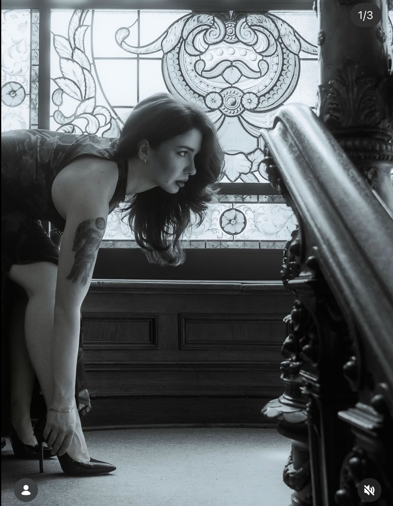 Czarno-białe zdjęcie kobiety w sukience, poprawiającej buta na tle zdobionego okna i balustrady schodów. Subtelne oświetlenie podkreśla detale.
