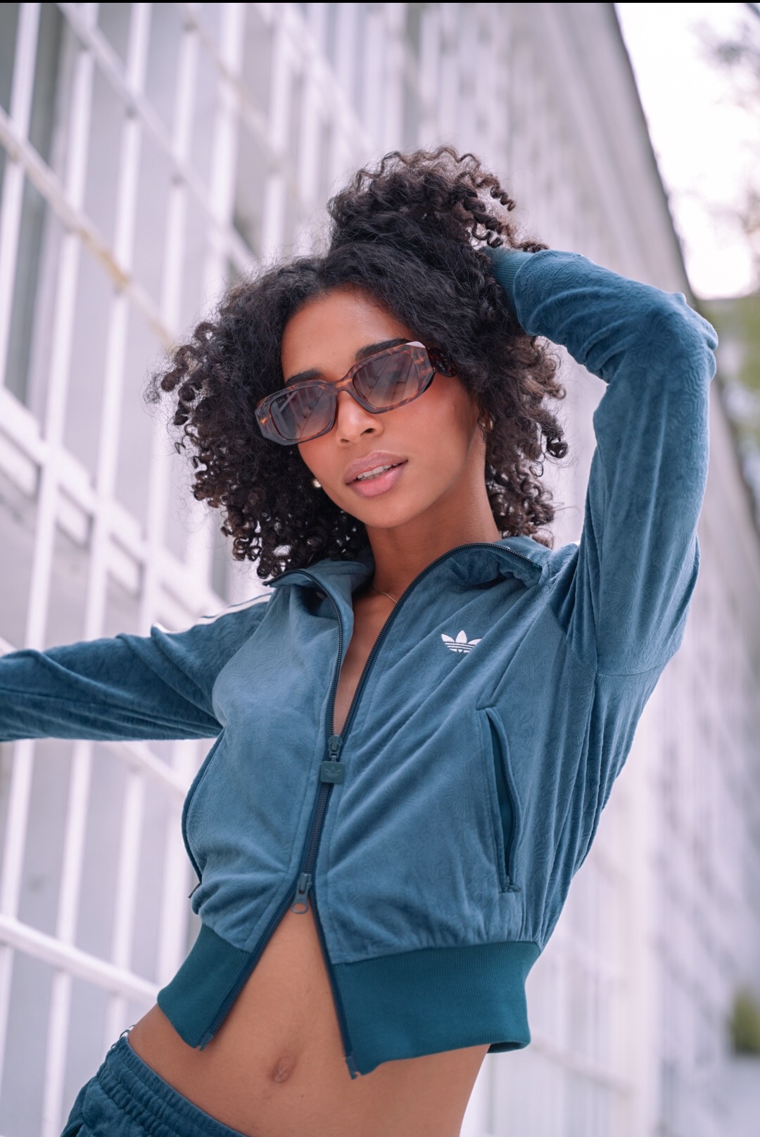 Portret kobiety w welurowym dresie Adidas, pozującej na tle białej ściany z kratownicą. Uwagę przyciągają brązowe okulary i kręcone włosy modelki.