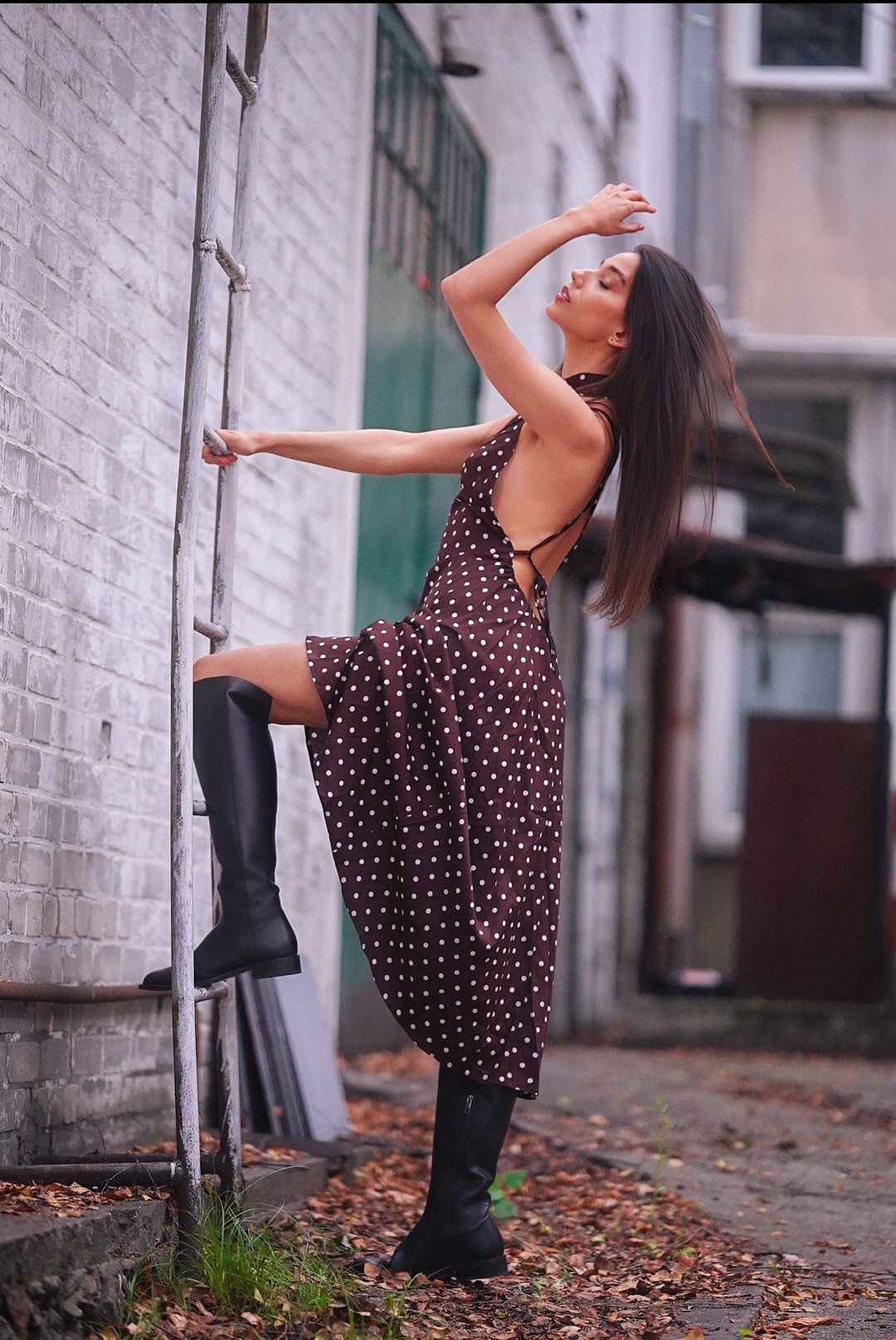 Kobieta w brązowej sukience w białe kropki i czarnych kozakach pozuje na tle ceglanej ściany, opierając się o metalową drabinę. Sesja w miejskiej scenerii.