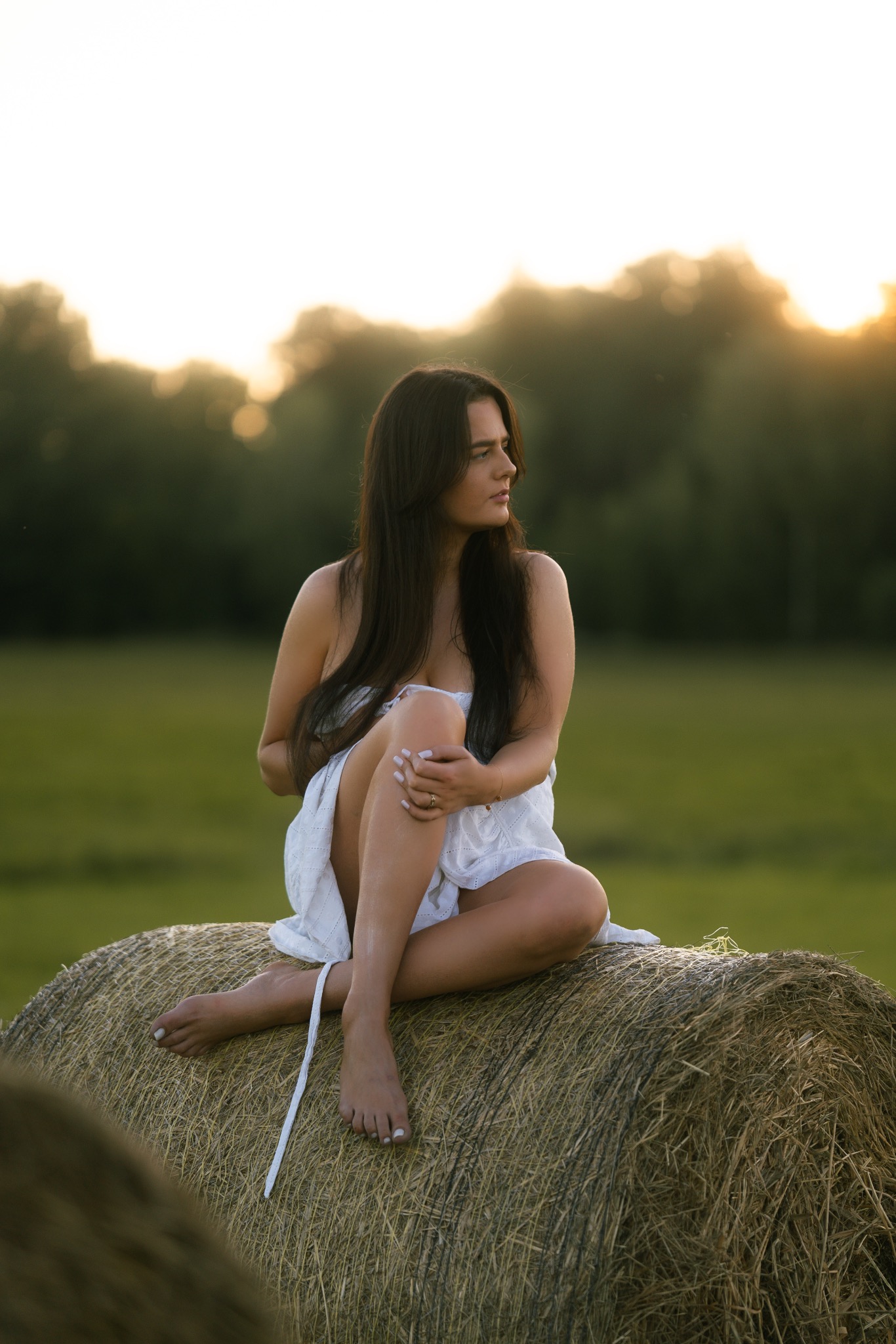 Kobieta w białej sukience siedzi na słomie na tle zielonego pola i drzew w promieniach zachodzącego słońca. Naturalna sesja w plenerze.
