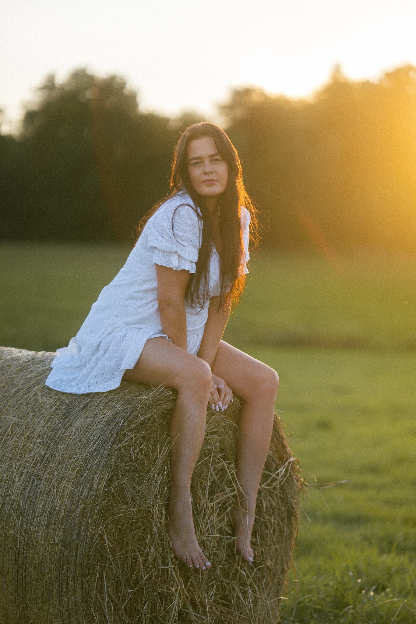 Kobieta w białej sukience siedzi boso na belocie siana w promieniach zachodzącego słońca. Romantyczna sceneria, naturalne światło, swobodna poza.