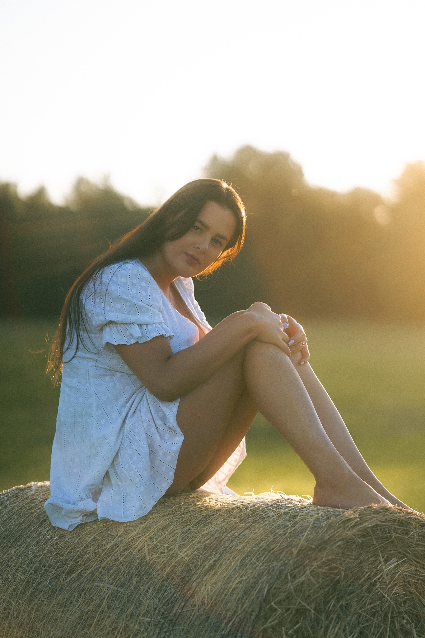 Portret kobiety w białej sukience siedzącej na belocie siana w promieniach zachodzącego słońca. Naturalne światło, miękkie tło z zielenią. Romantyczna sesja w plenerze.
