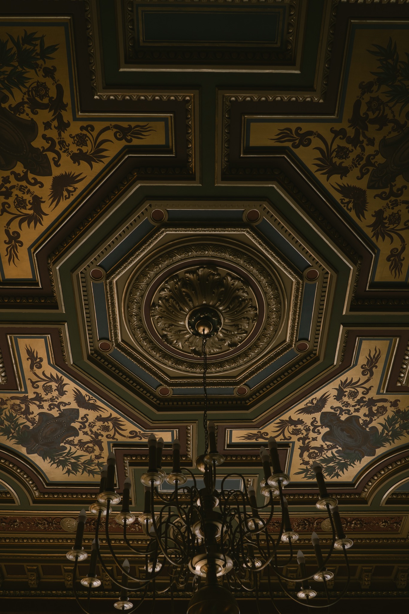 Dekoracyjny sufit z ornamentami w odcieniach złota i brązu, z żyrandolem w stylu vintage. Ujęcie z dołu podkreśla bogactwo detali i klasyczny charakter wnętrza.