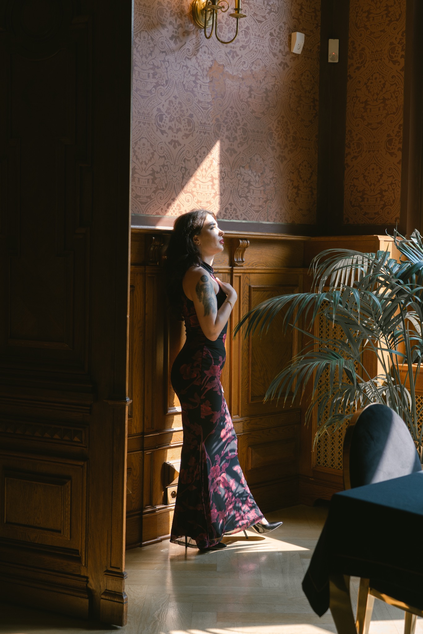 Elegancka kobieta w długiej sukni pozuje w promieniach słońca w stylowym wnętrzu z drewnianą boazerią i wzorzystą tapetą. Widoczny tatuaż na ramieniu i roślina doniczkowa.