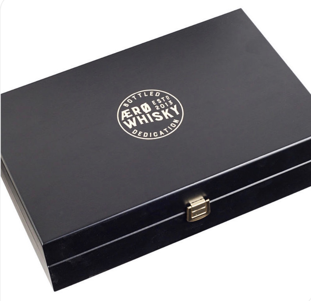 Czarna, lakierowana skrzynka z metalowym zapięciem, ozdobiona okrągłym, białym logo 'AERØ WHISKY DEDICATION' w prawym górnym rogu. Elegancki design, widoczny detal.