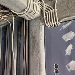 Jarosław Balaka - Instalacja elektryczna w trakcie montażu: liczne białe peszle z przewodami biegnące po surowym suficie i ścianie, z wejściem do płyty gipsowo-kartonowej z trzema otworami na puszki.