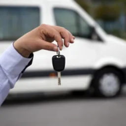 Dłoń w błękitnej koszuli trzyma kluczyk do białego samochodu dostawczego w tle.