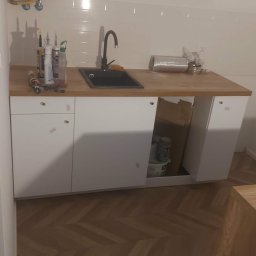 Nova-Bud - Nowoczesna zabudowa kuchenna z czarnym zlewem i drewnianym blatem, białe fronty szafek, parkiet w jodełkę na podłodze. Widoczne narzędzia i materiały budowlane.