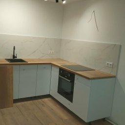 Nova-Bud - Nowoczesna kuchnia z białymi szafkami, drewnianym blatem i marmurowym panelem ściennym. Czarny zlew i płyta indukcyjna. Minimalistyczny design, jasne wnętrze.