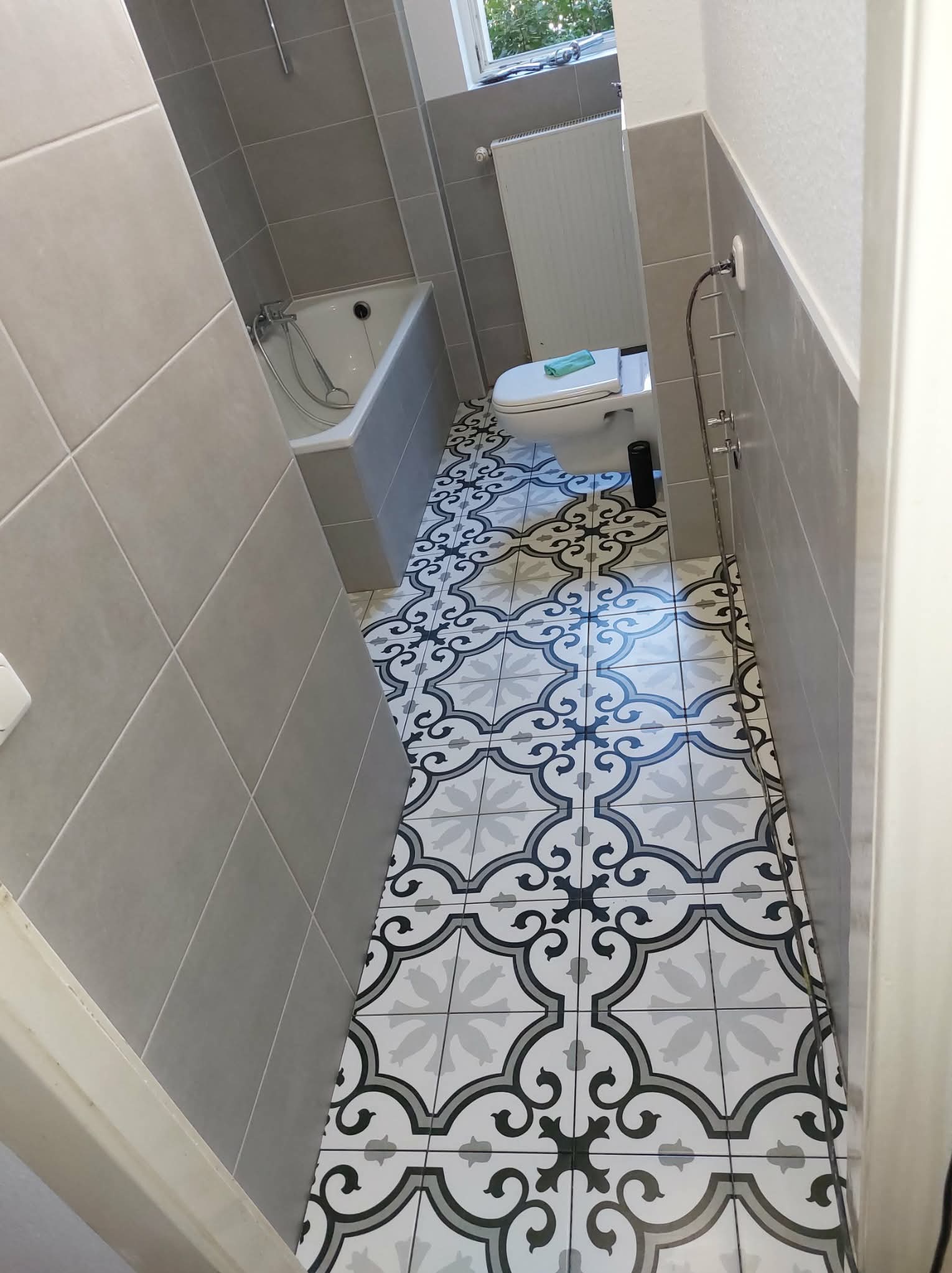 Wykończona łazienka z szarymi płytkami ściennymi i wzorzystą podłogą w stylu retro. Widoczna wanna, toaleta i grzejnik. Ujęcie z perspektywy wejścia.