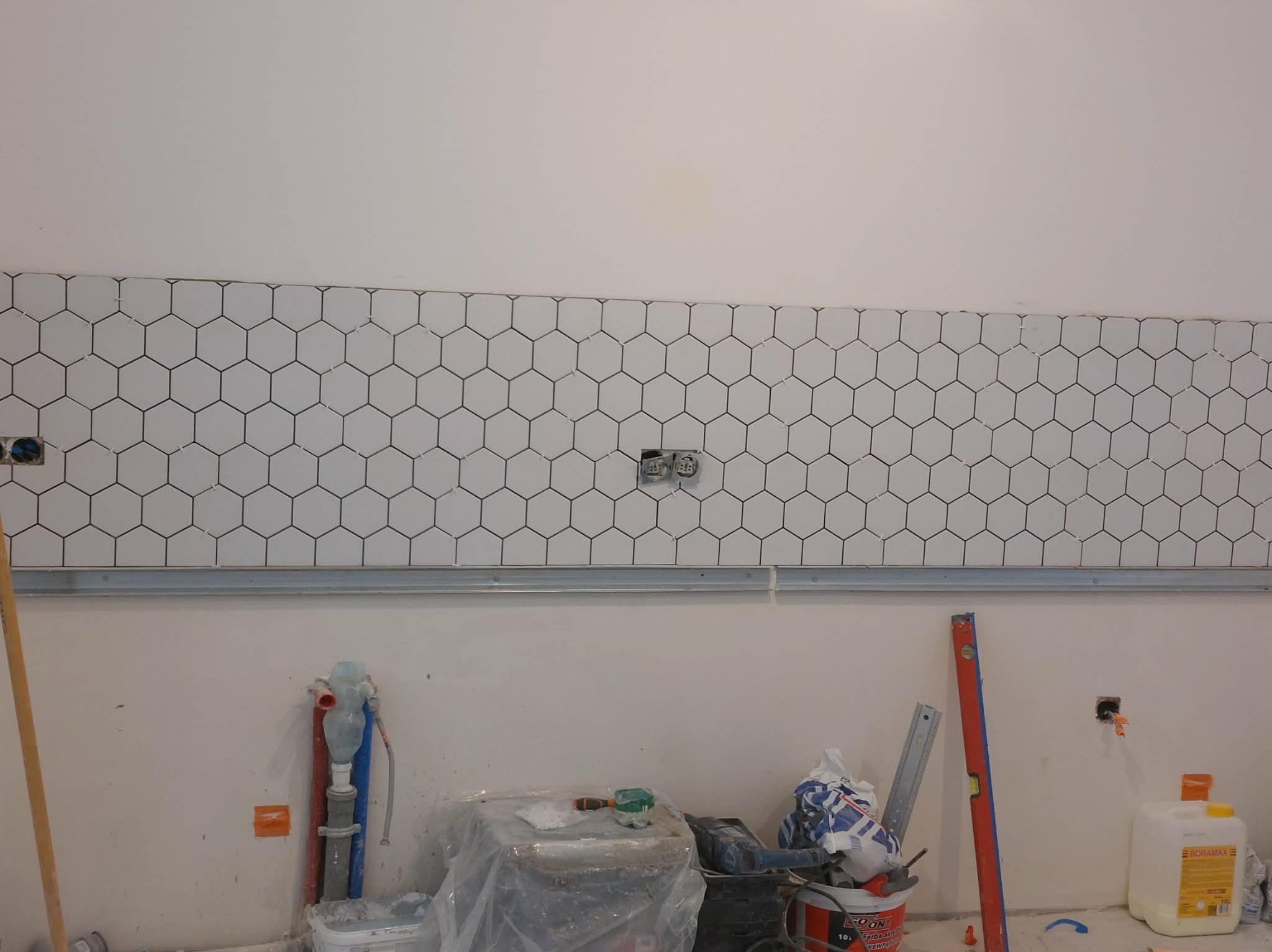 Układanie heksagonalnych płytek na ścianie w trakcie remontu. Widoczne narzędzia glazurnicze i puszki elektryczne. Płytki białe z czarną fugą, zamontowane na metalowej listwie.