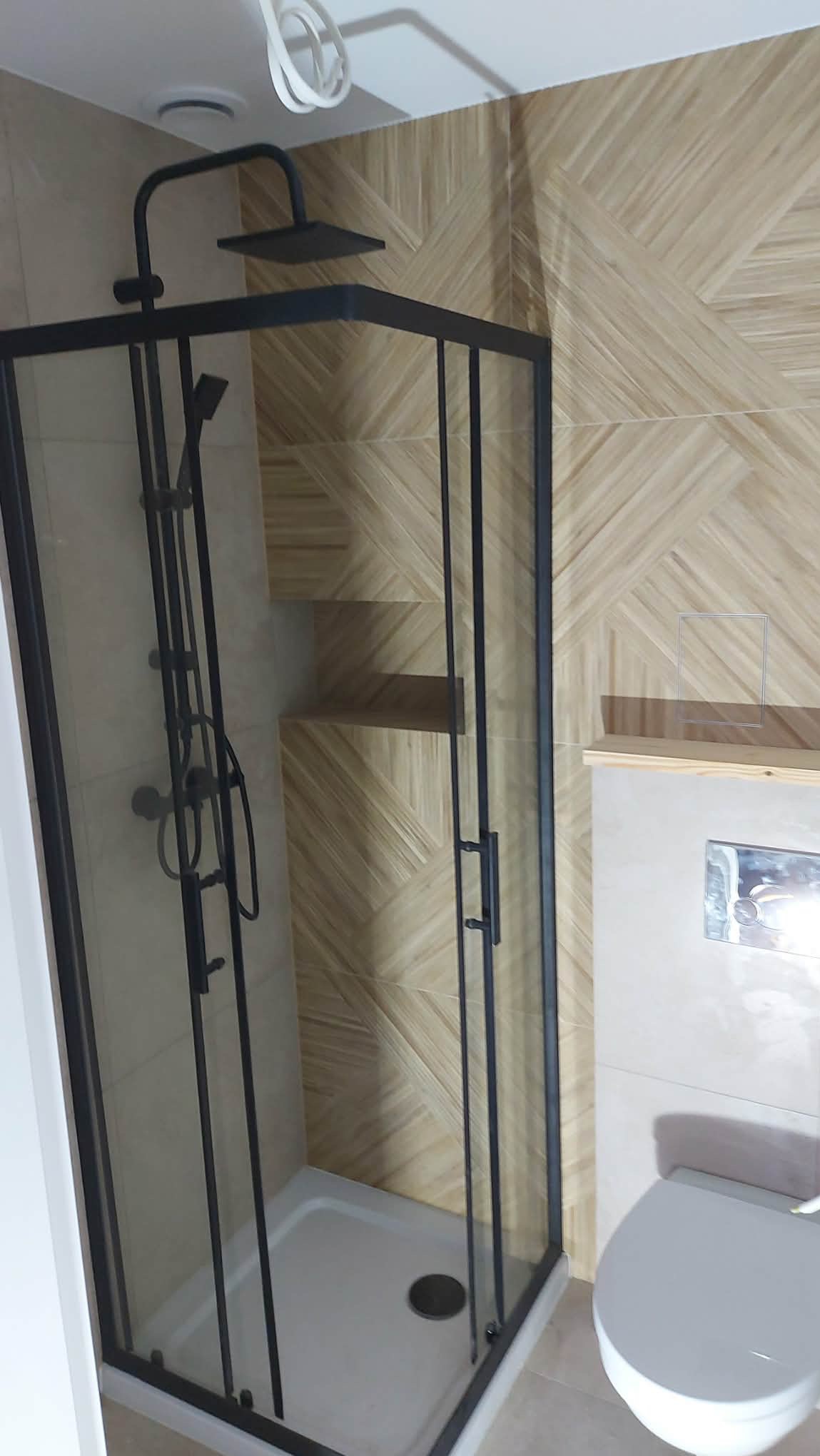 Nowoczesna kabina prysznicowa z czarnymi profilami na tle beżowych płytek z geometrycznym wzorem. Widoczna toaleta i półka wewnątrz kabiny. Wykończenie łazienki.