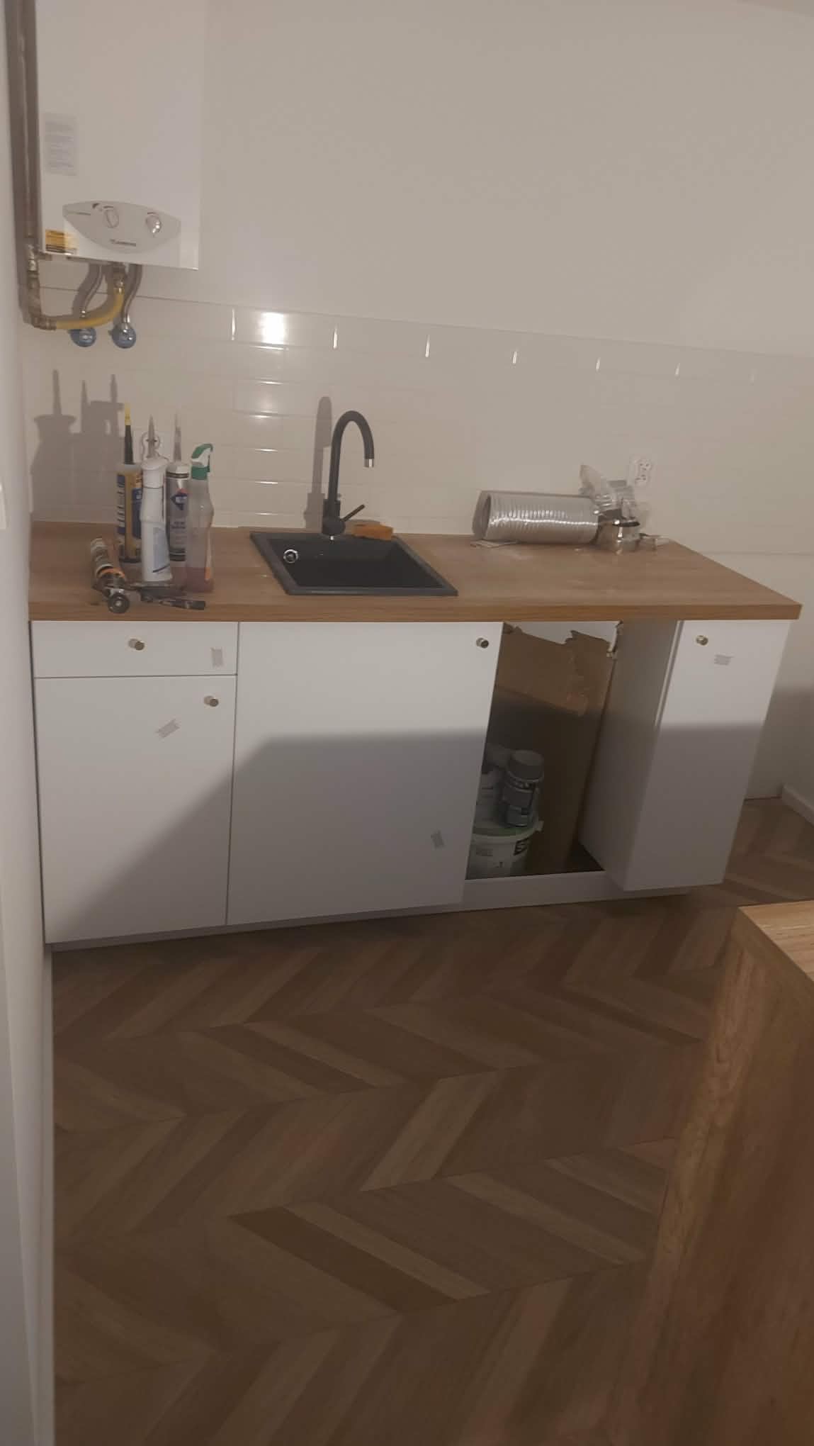 Nowoczesna zabudowa kuchenna z czarnym zlewem i drewnianym blatem, białe fronty szafek, parkiet w jodełkę na podłodze. Widoczne narzędzia i materiały budowlane.